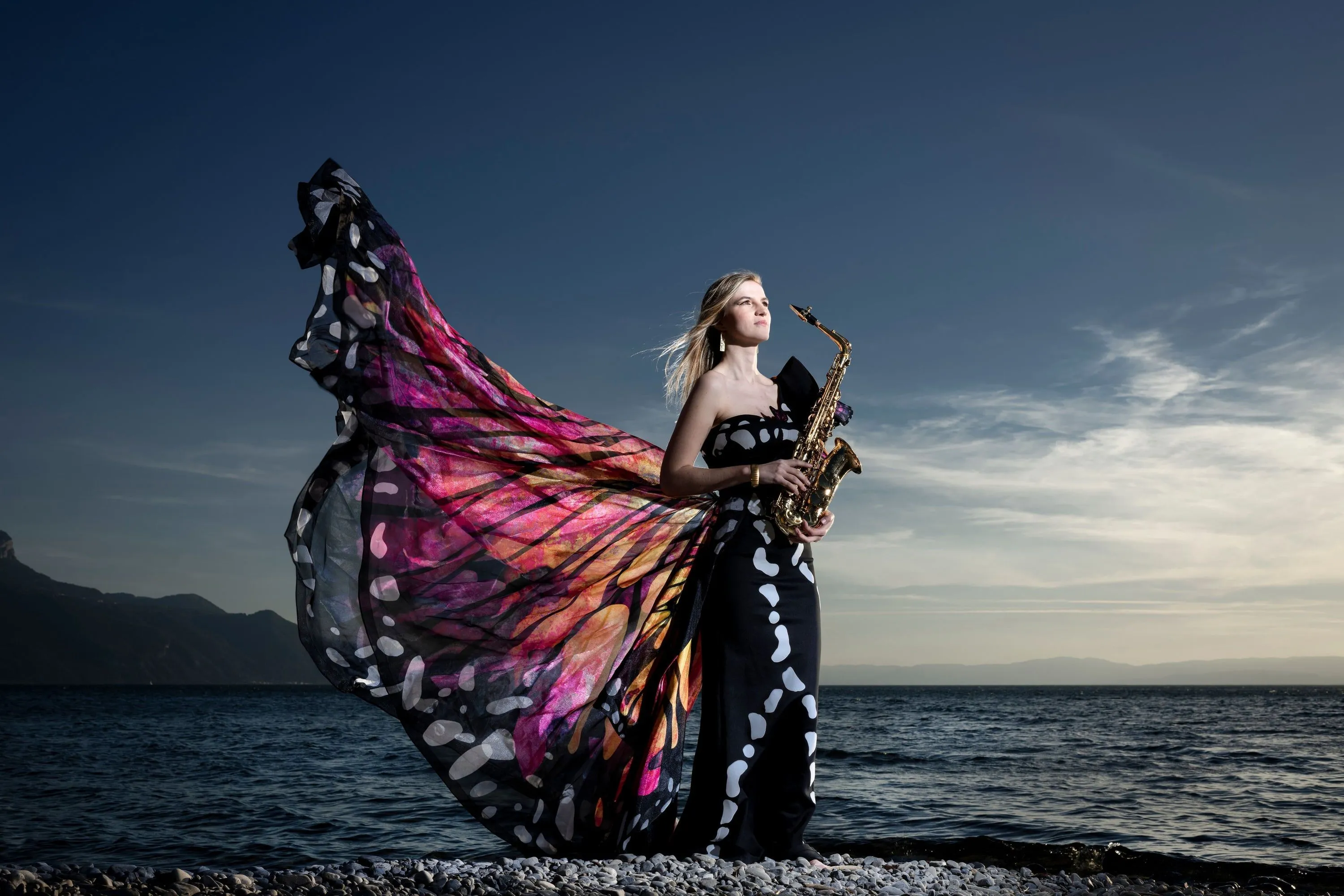 Valentine Michaud with a saxophone on a lakeshore at dusk, wearing large butterfly wings